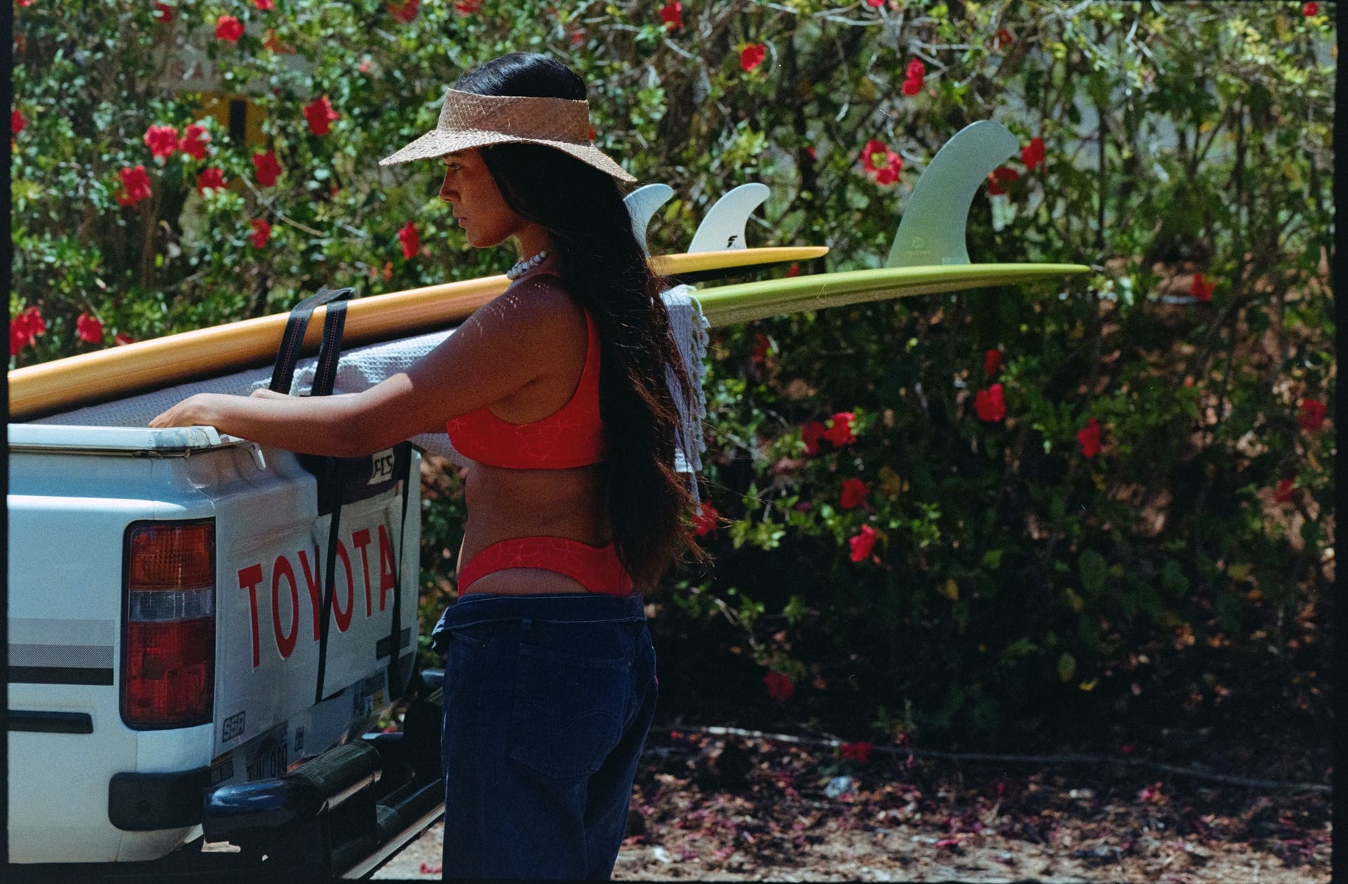 girl in red bikini and straw hat hanging on the back of the truck bed with surfboards in the back strapping in
