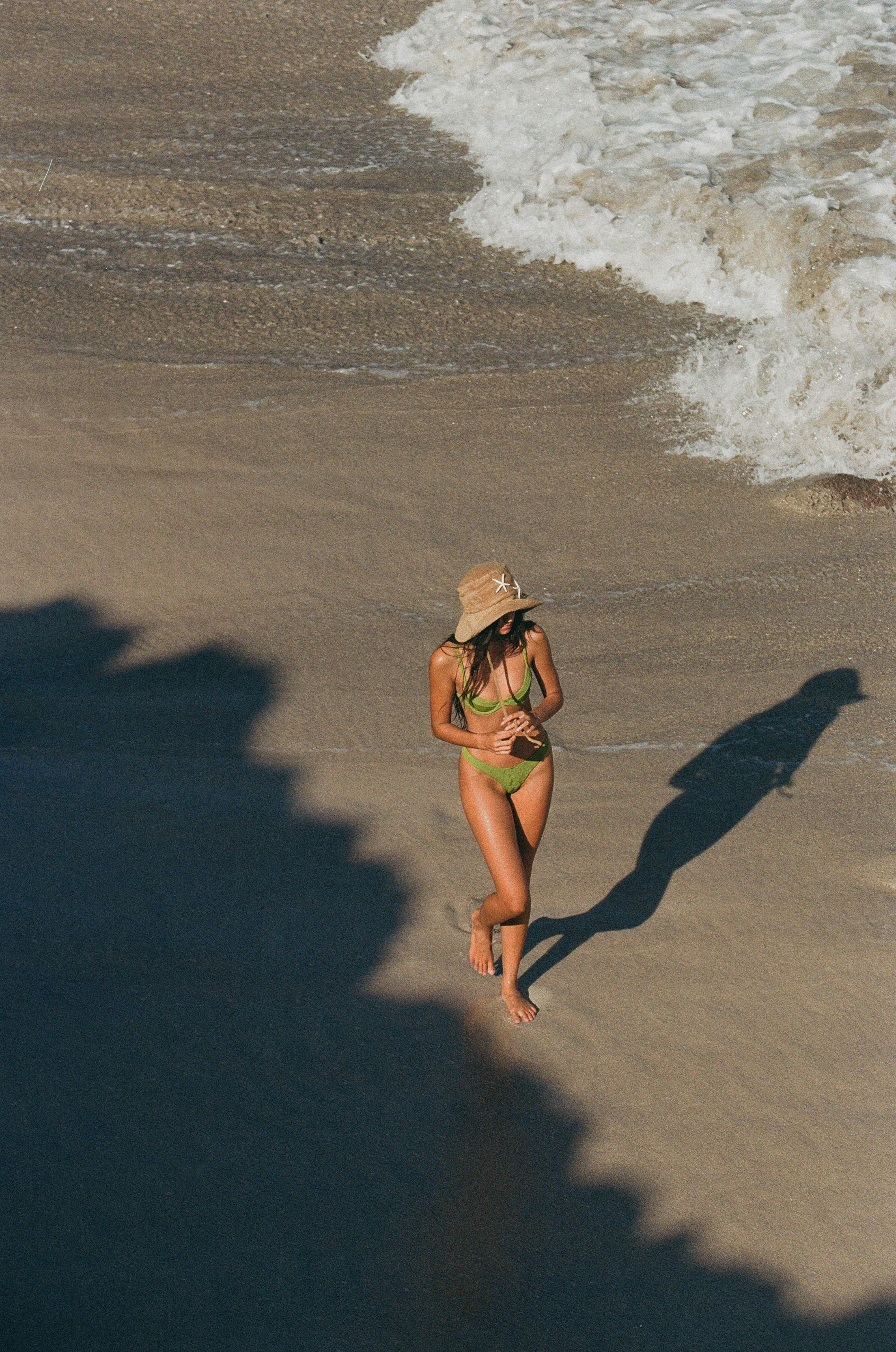 girl walking long shoreline in eige hat in a green bikini