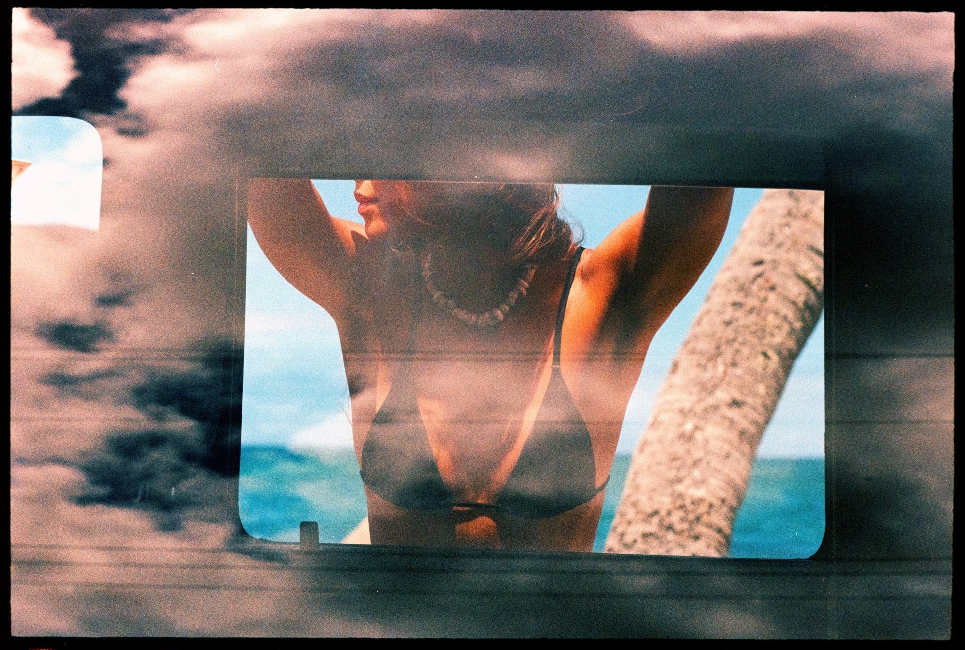 girl leaning with arms up on the side of the truck in a bikini with a shell necklace checking the ocean, looking through the window of the truck